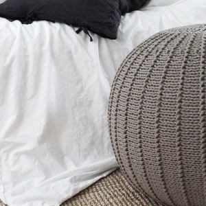 May include: A grey knitted pouffe ottoman sits on a natural fibre rug in front of a white bed with a black pillow.