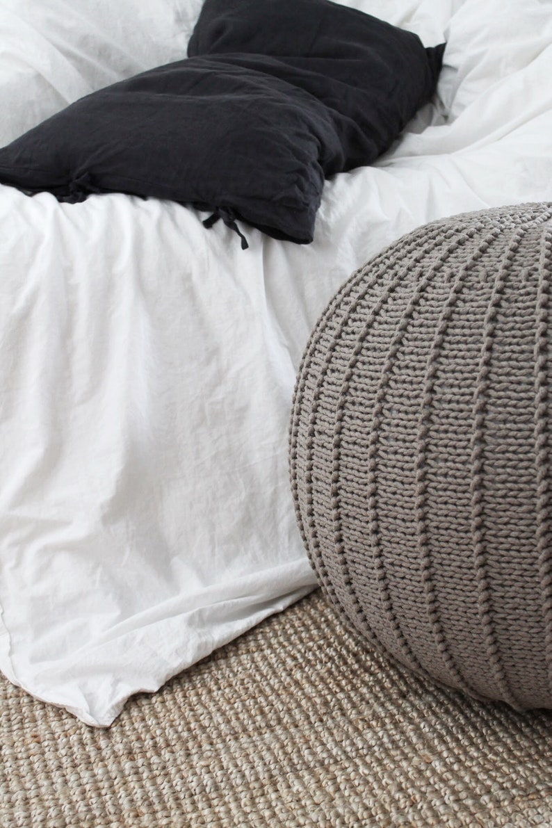 May include: A grey knitted pouffe ottoman sits on a natural fibre rug in front of a white bed with a black pillow.