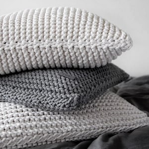 May include: A stack of three knitted throw pillows. The top pillow is white, the middle pillow is gray, and the bottom pillow is white. The pillows are arranged on a gray fabric surface.