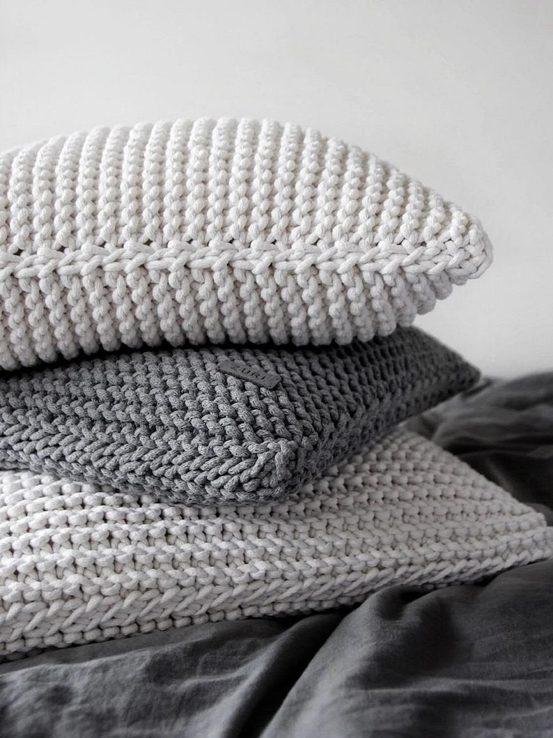 May include: A stack of three knitted throw pillows. The top pillow is white, the middle pillow is gray, and the bottom pillow is white. The pillows are arranged on a gray fabric surface.