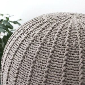 May include: A large, round, knitted pouffe in a light brown colour. The pouffe is made of thick, chunky yarn and has a textured surface.