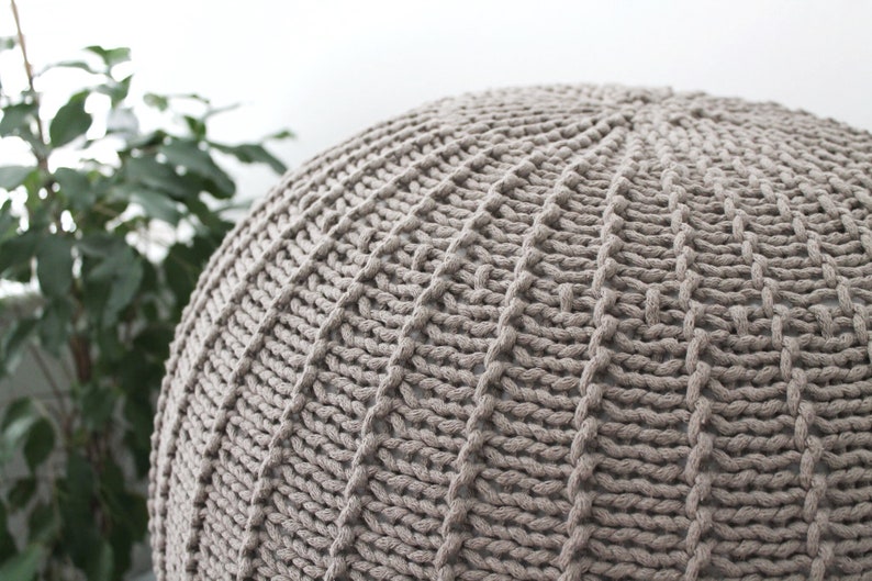 May include: A large, round, knitted pouffe in a light brown colour. The pouffe is made of thick, chunky yarn and has a textured surface.