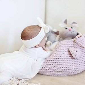 Puede incluir: Un beb&eacute; con una diadema blanca con un lazo, acostado en el suelo junto a una cesta de crochet rosa con un juguete de crochet de conejo en su interior.