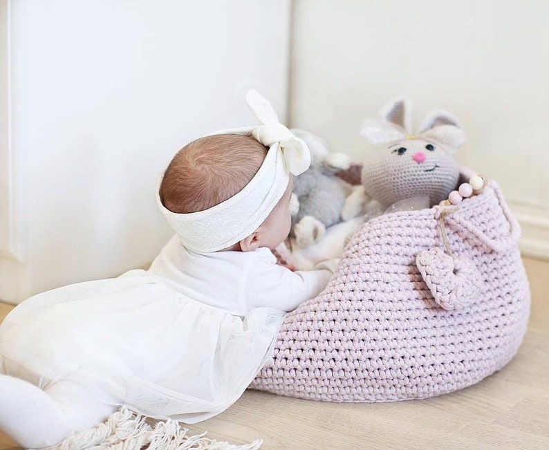 Puede incluir: Un beb&eacute; con una diadema blanca con un lazo, acostado en el suelo junto a una cesta de crochet rosa con un juguete de crochet de conejo en su interior.