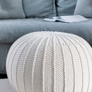 May include: A white knitted round ottoman with a textured surface. The ottoman is sitting on a white rug.