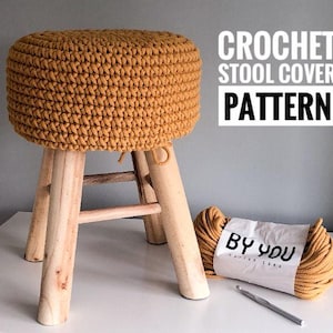 May include: A crocheted stool cover in a mustard yellow color. The cover is made with thick yarn and has a simple, textured design. The stool cover is sitting on a wooden stool with four legs. A ball of yarn and a crochet hook are also in the image. The yarn label reads "By You Cotton Cord".