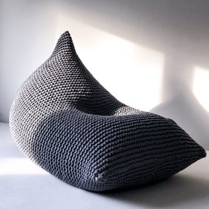May include: A grey knitted bean bag chair with a unique teardrop shape. The chair is made of thick, chunky yarn and has a soft, comfortable texture.