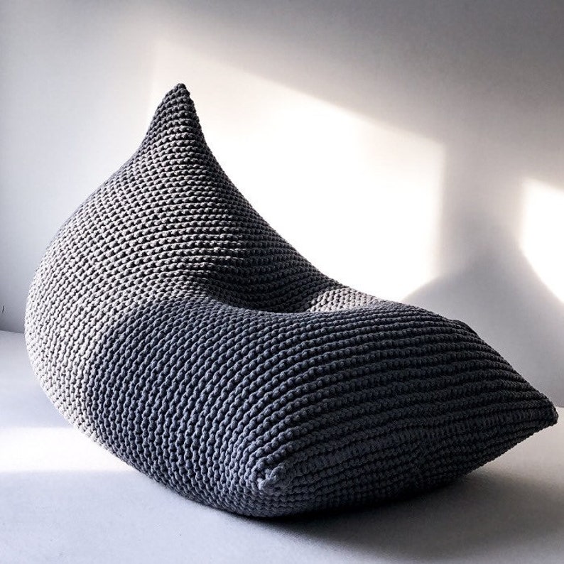 May include: A grey knitted bean bag chair with a unique teardrop shape. The chair is made of thick, chunky yarn and has a soft, comfortable texture.
