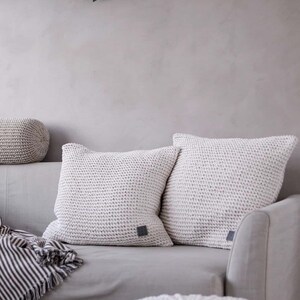 May include: Two white knitted throw pillows with a subtle texture on a gray couch. The pillows have a small gray square label on each. A white knitted pouf sits in the foreground.