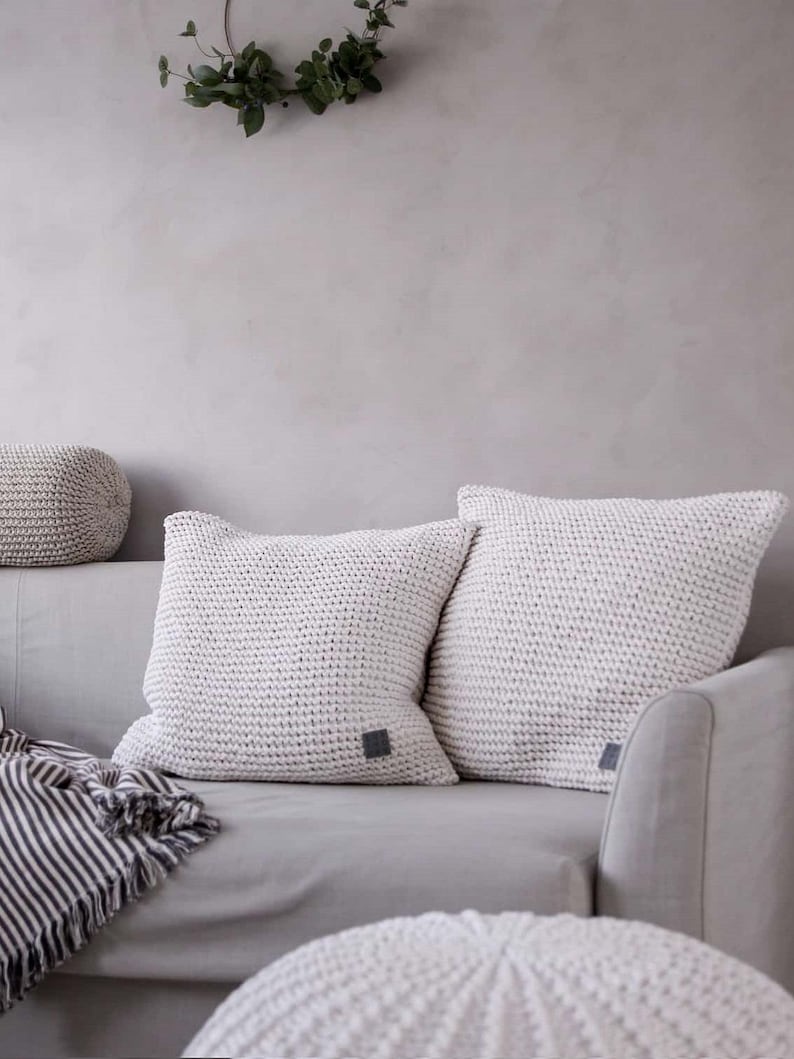 May include: Two white knitted throw pillows with a subtle texture on a gray couch. The pillows have a small gray square label on each. A white knitted pouf sits in the foreground.