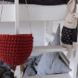 May include: A red crocheted hanging basket with a white cord. The basket is hanging from a white shelf. A crocheted bunny toy is on the shelf.