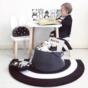 Puede incluir: Un conjunto de mesa y silla blanca con una alfombra rayada en blanco y negro. La silla tiene orejas de conejo y un patr&oacute;n de estrellas blancas y negras en el asiento. Un ni&ntilde;o est&aacute; sentado en la mesa leyendo un libro. Hay animales de peluche y un libro titulado "Black Cat White Cat" en una cesta gris.