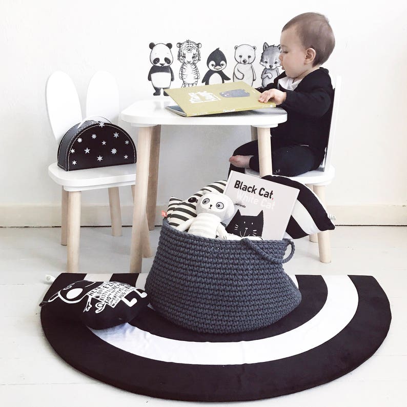 Puede incluir: Un conjunto de mesa y silla blanca con una alfombra rayada en blanco y negro. La silla tiene orejas de conejo y un patr&oacute;n de estrellas blancas y negras en el asiento. Un ni&ntilde;o est&aacute; sentado en la mesa leyendo un libro. Hay animales de peluche y un libro titulado "Black Cat White Cat" en una cesta gris.