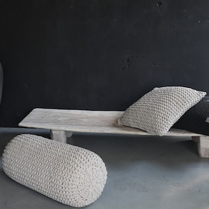 May include: A light gray knitted bolster pillow sits on a gray floor in front of a wooden bench. Two knitted pillows, one light gray and one black, are on the bench. A large, dark gray ceramic pot is in the background.