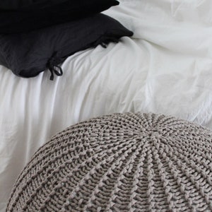 May include: A large, round, knitted pouffe in a light brown colour. The pouffe is made of thick, textured yarn and has a circular pattern.