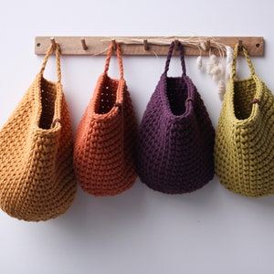 May include: Four crocheted hanging baskets in different colors: mustard yellow, burnt orange, purple, and olive green. The baskets are hanging from a wooden rack.