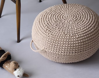 May include: A beige crocheted round pouf with a handle. The pouf is made of thick yarn and has a textured surface.