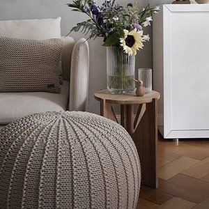 May include: A beige knitted pouffe ottoman with a light beige knitted throw pillow on a white sofa. A vase with a sunflower and other flowers sits on a wooden side table.