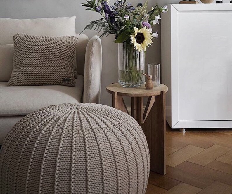 May include: A beige knitted pouffe ottoman with a light beige knitted throw pillow on a white sofa. A vase with a sunflower and other flowers sits on a wooden side table.