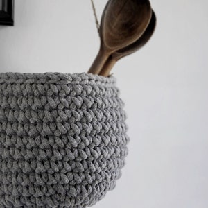 May include: A gray crocheted hanging basket with two wooden spoons inside. The basket is hanging from a white wall.