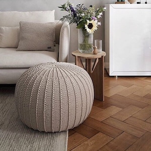 May include: A beige knitted ottoman with a round shape and a textured surface. The ottoman is sitting on a light beige rug in front of a white sofa.