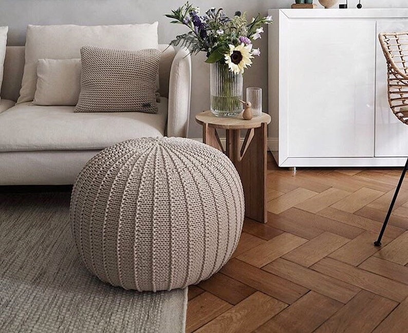 May include: A beige knitted ottoman with a round shape and a textured surface. The ottoman is sitting on a light beige rug in front of a white sofa.