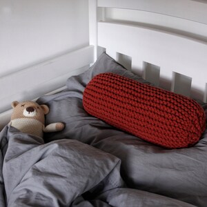May include: A red knitted cylindrical pillow rests on a gray bedspread. A brown crocheted teddy bear is lying on the bedspread.