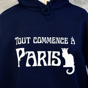 Könnte beinhalten: Ein marineblauer Hoodie mit einem weißen Grafikdruck, der "Tout commence à Paris" mit einer weißen Silhouette einer Katze neben dem Text sagt.