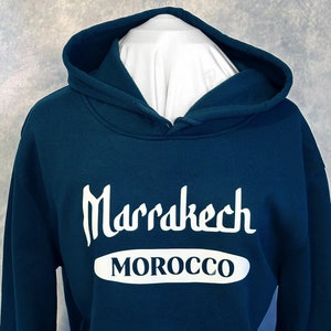 Könnte beinhalten: Ein dunkel blaugrüner Kapuzenpullover mit weißem Text, der "Marrakech MOROCCO" lautet.