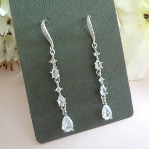 May include: A pair of silver-toned drop earrings featuring a curved hook set with small clear stones, followed by a vertical chain of alternating round and pear-shaped clear faceted stones that dangle downward in a graduated, elegant arrangement.