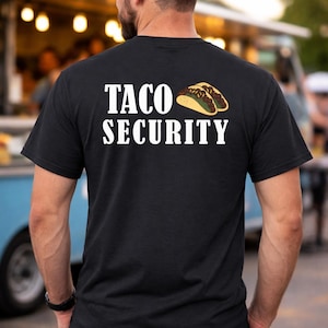 May include: Black t-shirt with the words "TACO SECURITY" in white block letters and a cartoon taco graphic. The person is wearing blue jeans and a black watch. The background is a food truck.