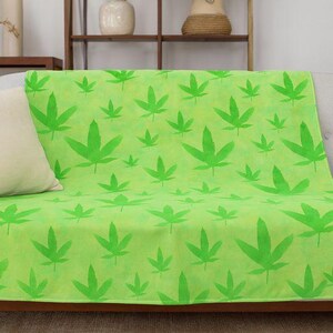 May include: A lime green throw blanket with a repeating pattern of bright green cannabis leaves. The blanket is draped over a light gray sofa, with a cream-colored pillow on the left.