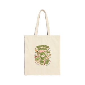 May include: A beige canvas tote bag featuring a central graphic with the text "MATCHA RITUAL". The illustration includes a small character, a bamboo whisk, a matcha bowl, a jar, a cup, and a drink, surrounded by small hearts and strawberries.