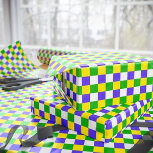 May include: Two gift boxes wrapped in colorful checkered paper in green, yellow, white, and purple. A roll of matching wrapping paper and a pair of scissors are visible. Black ribbon is in the foreground. The scene is set near a window.