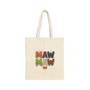 May include: A beige canvas tote bag featuring the text MAW MAW in a multi-colored Halloween-themed font. The letters are decorated with orange, purple, green, mummy-wrap, and black diamond patterns, accented by a small orange bow at the bottom.