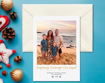 May include: A holiday card with a family photo on a beach, with the text "Dashing through the sand." The card is accompanied by a white envelope and festive decorations, including gold ornaments and a red felt star.