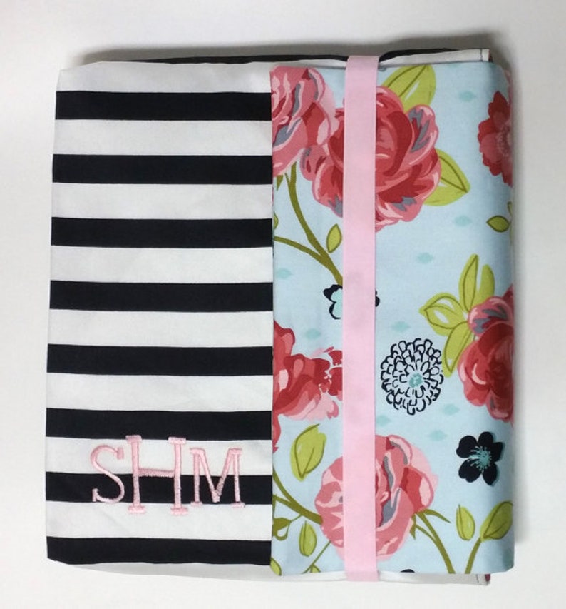 May include: A black and white striped fabric with a floral fabric panel sewn to it. The floral fabric has pink, blue, and green flowers on a light blue background. The striped fabric has the letters "SHM" embroidered on it in pink thread.