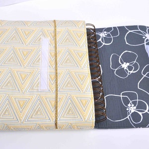 May include: A gray and white floral patterned notebook with a yellow and white geometric patterned cover. The notebook has a spiral binding and a white velcro closure.