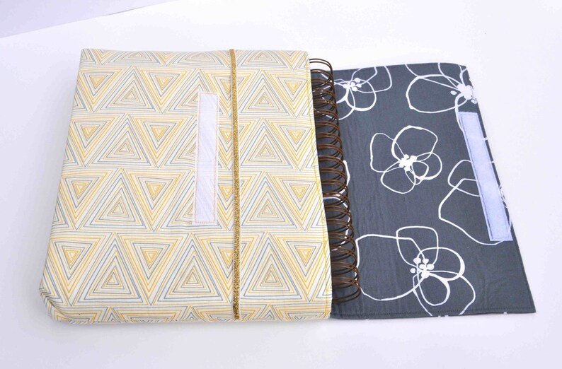 May include: A gray and white floral patterned notebook with a yellow and white geometric patterned cover. The notebook has a spiral binding and a white velcro closure.