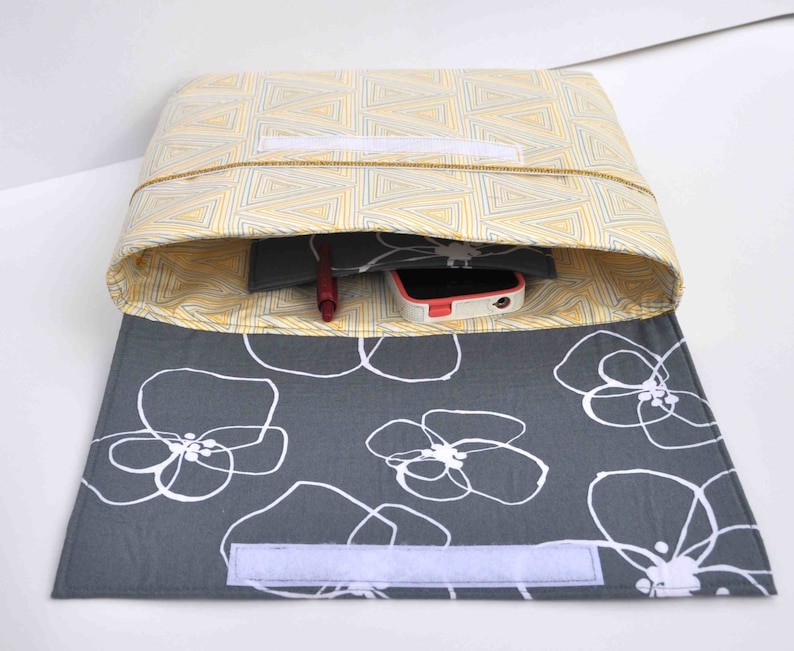 May include: A gray and yellow fabric pouch with a white zipper. The pouch is open and has a red pen, a black notebook, and a white phone inside. The pouch has a gray fabric lining with a white floral pattern.