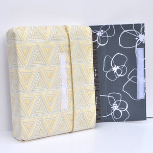May include: Two fabric-covered notebooks, one with a yellow and gray geometric pattern and the other with a gray background and white floral design. Both notebooks have a white velcro closure.