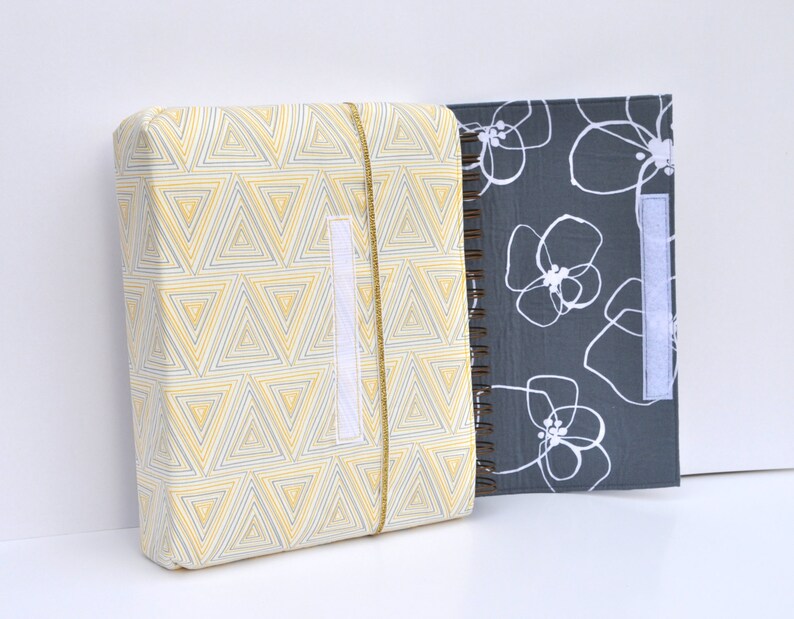 May include: Two fabric-covered notebooks, one with a yellow and gray geometric pattern and the other with a gray background and white floral design. Both notebooks have a white velcro closure.