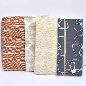 May include: Three fabric covered notebooks with different patterns. The first notebook has a brown and white chevron pattern. The second notebook has a white background with a brown leaf pattern. The third notebook has a gray background with a white floral pattern.