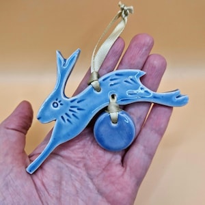 Puede incluir: Adorno de cerámica azul claro con forma de liebre, con una cinta dorada para colgar. La liebre está en posición de carrera con detalles grabados. Un pequeño círculo azul está unido a la liebre. El adorno se sostiene en una mano sobre un fondo melocotón.