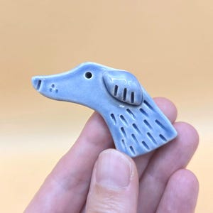 May include: A light blue ceramic brooch shaped like the head of a dog in profile. The piece features a glossy finish, a small circular eye, and incised vertical lines along the neck and ear area to provide texture and detail to the canine form.