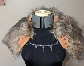May include: A faux fur shoulder piece with brown and gray tones, featuring leather accents and a silver chain with decorative spikes. The piece is displayed on a black dress form.