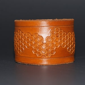 May include: A brown leather cuff bracelet with a honeycomb pattern. The cuff is a warm, medium brown color and features a repeating hexagonal design. The bracelet is a wide band, likely designed to be worn on the wrist.