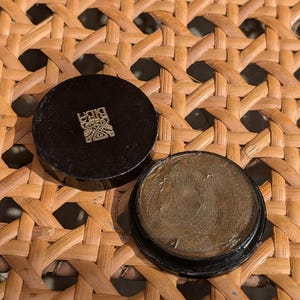 Könnte beinhalten: Ein kleiner runder schwarzer Behälter mit einem goldenen Design auf dem Deckel. Der Behälter ist geöffnet und eine goldfarbene Substanz ist im Inneren sichtbar.