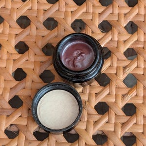 May include: An open, black jar of dark red-brown cosmetic product, with the lid beside it. The product is on a woven, light brown surface.