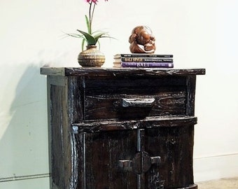 May include: A dark wood cabinet with a drawer and two doors. The cabinet has a distressed finish and is decorated with metal accents. A small wooden statue, a vase with pink flowers, and a stack of books are on top of the cabinet.